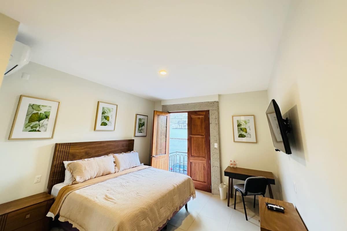 Un dormitorio con cama, escritorio y televisión. Hay cuadros en la pared y una puerta que da al exterior.
