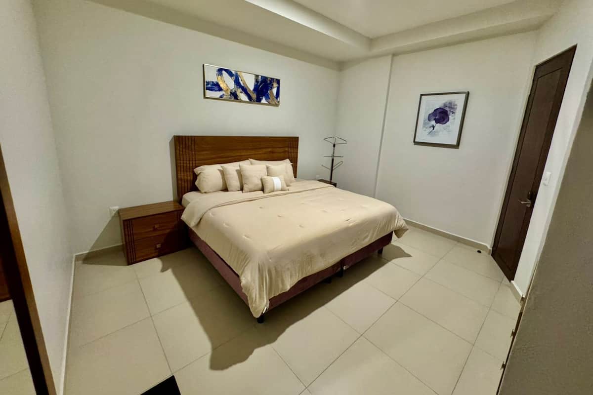 Un dormitorio con cama, mesita de noche, lámpara de pie y dos cuadros colgados en la pared.