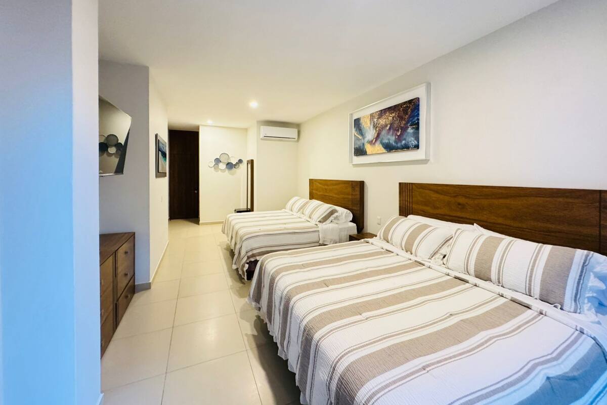 Habitación de hotel con dos camas, cabecera de madera, un cuadro en la pared y un pasillo que conduce a otra habitación.