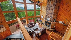 A cozy cabin interior with large windows, a stone fireplace, and a wooden deck.