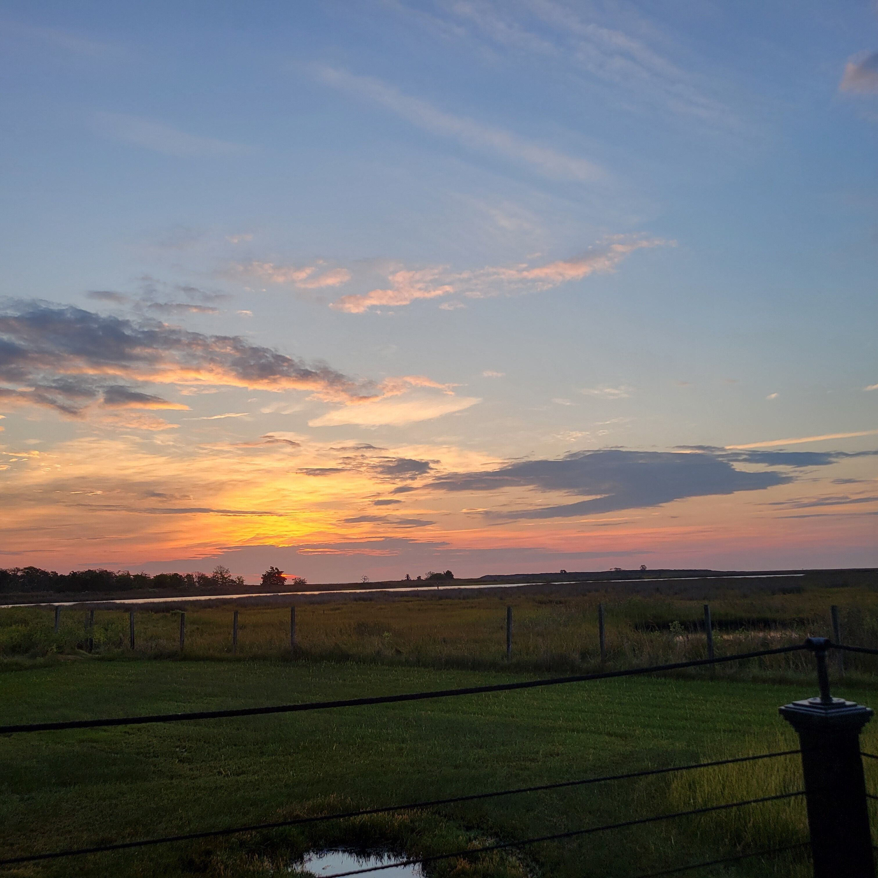 Een zonsondergang boven een grasveld met een hek en een lantaarnpaal.