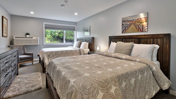 A bedroom with two beds, a dresser, a window with a view of greenery, and a painting on the wall.