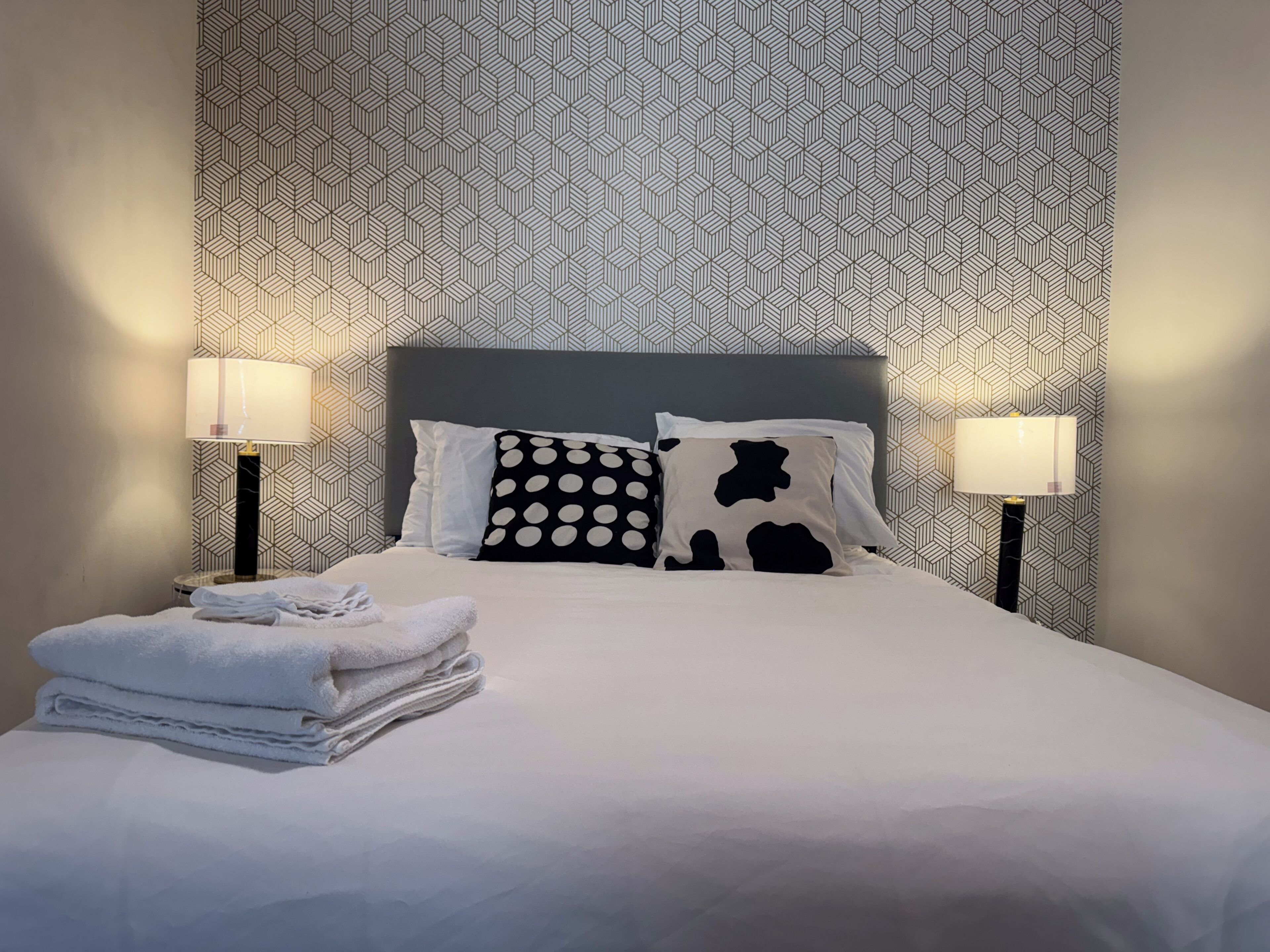 A neatly made bed with white linens and two black and white patterned pillows, flanked by two bedside lamps on either side.