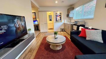 A modern living room with a flat-screen TV, a round coffee table, a black sofa with red and grey cushions, and a kitchen area with a microwave and a sink.