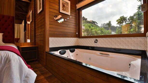 A modern bathroom with a large bathtub, wooden walls, and a window offering a view of greenery. - Stillo Gramado Dutra (Gramado)