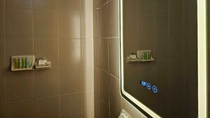 Une salle de bain carrelée de carreaux beiges, dotée d’une douche en verre et de deux petites étagères où sont rangés les articles de toilette.