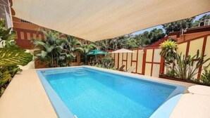 Une piscine rectangulaire à l’eau d’un bleu cristallin, entourée de palmiers et dotée d’un espace ombragé sous une pergola.