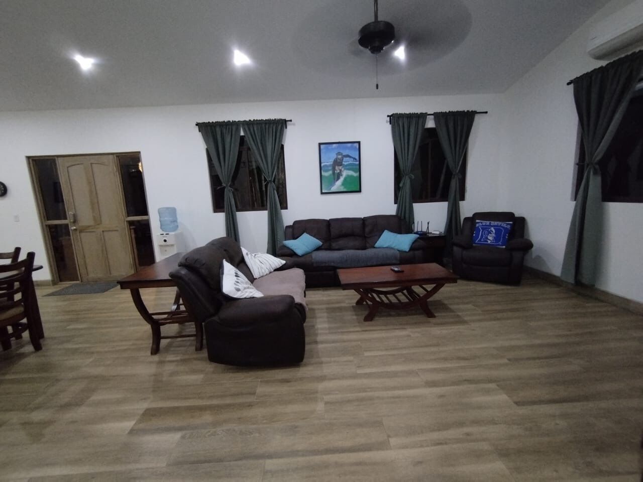 A spacious living room with a sofa, armchair, and coffee table. There is a door and a window with curtains.
