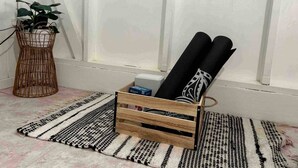 A room with a wooden crate holding rolled-up mats, a wicker plant stand, and a striped rug on the floor.