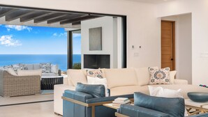 Ein modernes Wohnzimmer mit einer Couch, Sesseln und einem Couchtisch. Ein großes Fenster bietet Blick auf den Ozean.