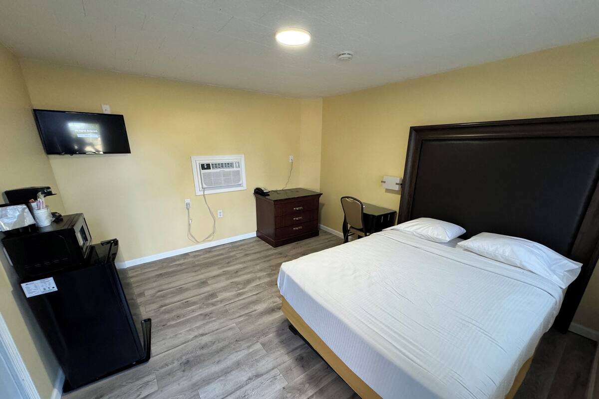 A hotel room with a bed, a desk, a dresser, a television, and an air conditioning unit.