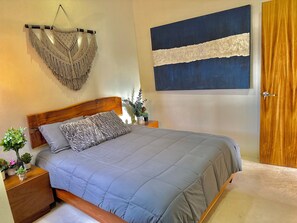Una camera da letto con un letto, una testiera in legno, comodini, un arazzo in macramè, un quadro blu e una porta in legno.