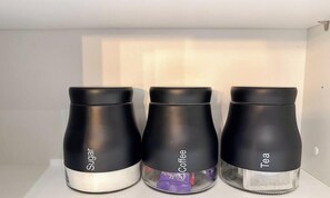 Três recipientes pretos identificados como 'Sugar', 'Coffee' e 'Tea' sobre uma prateleira branca.