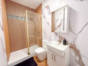 A modern bathroom with a glass-enclosed shower, a white toilet, and a marble-patterned wall. - Modern 2 Bedroom Flat in Luton Town Centre (Luton)