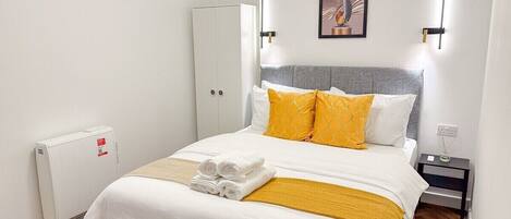 Una cama bien tendida con ropa de cama blanca y amarilla, un cabecero gris y un radiador blanco.