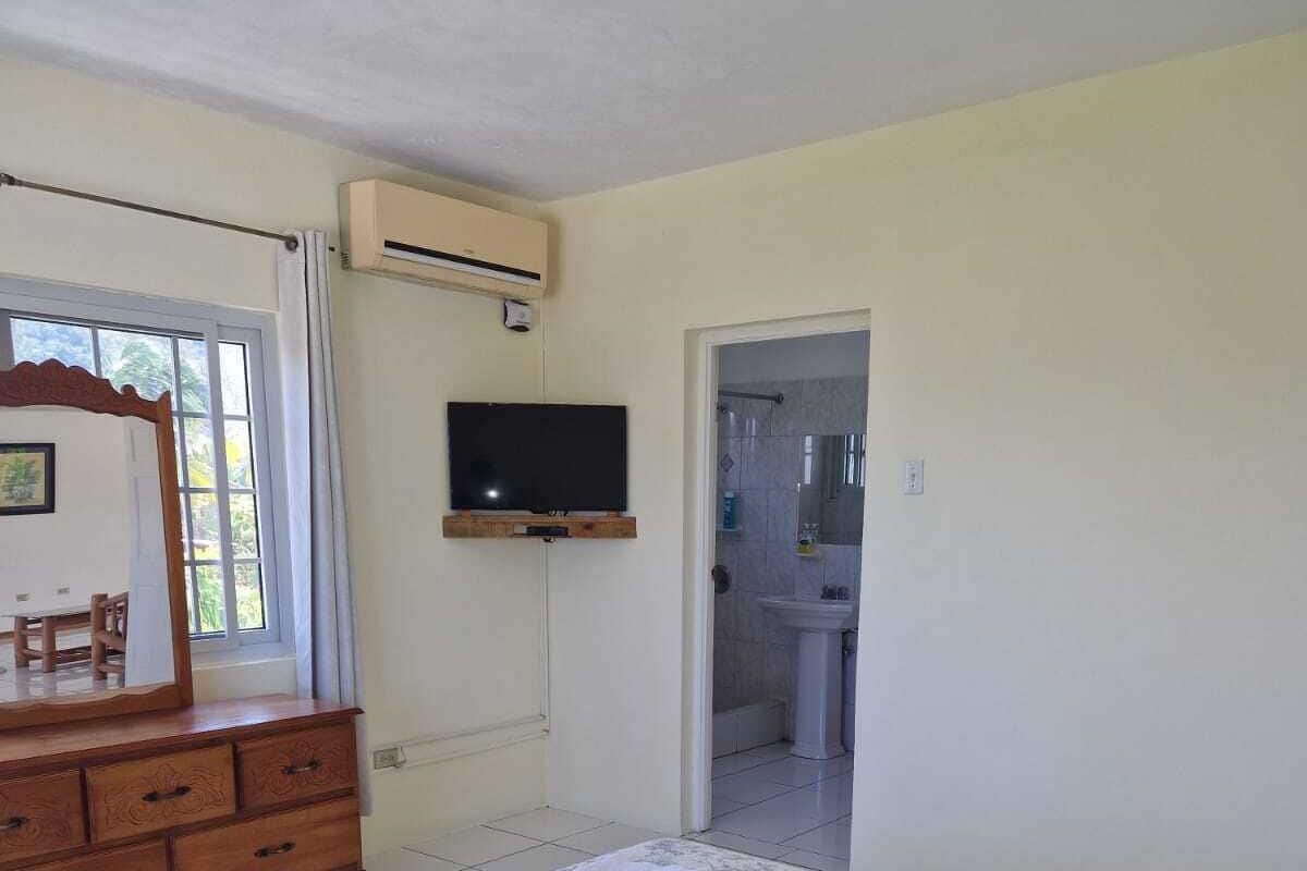 A bedroom with a bed, a wooden dresser, a television, and a bathroom visible through an open door.