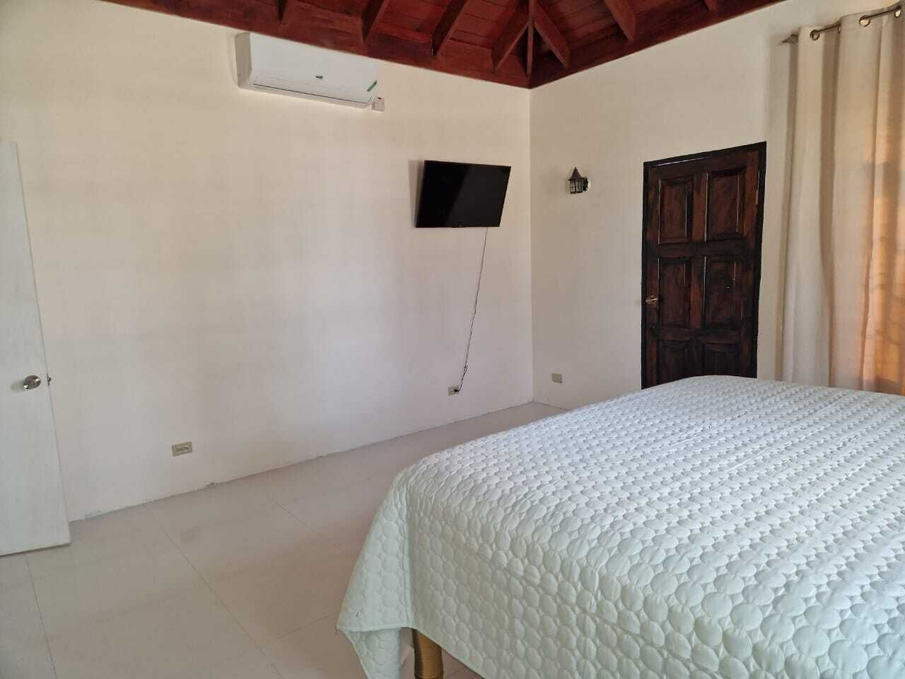 A bedroom with a bed, a television mounted on the wall, a wooden door, and a window with curtains.
