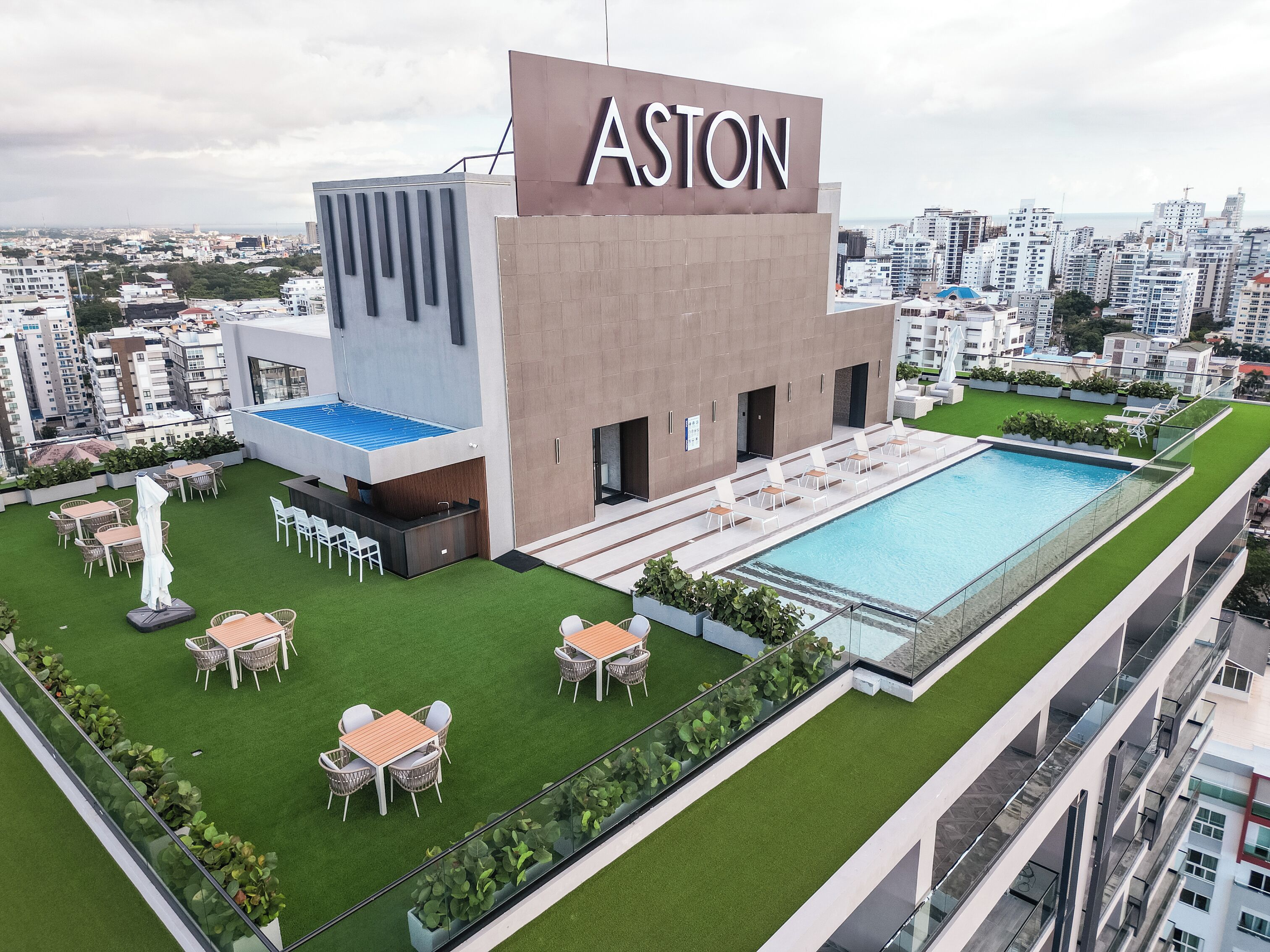 A modern building with the name "ASTON" on the facade, a rooftop pool, and outdoor seating areas.