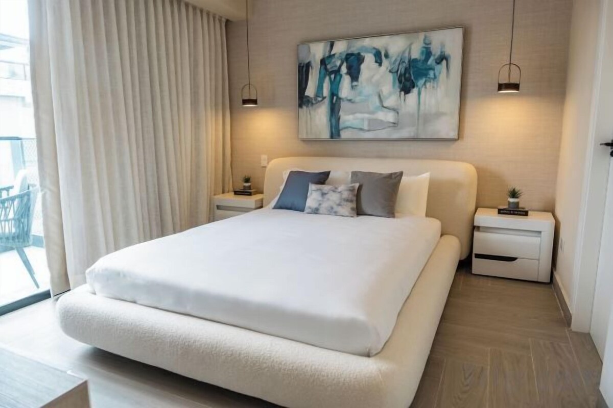 Habitación de hotel con una cama grande, una mesita de noche con lámpara, un cuadro en la pared y vistas al exterior.