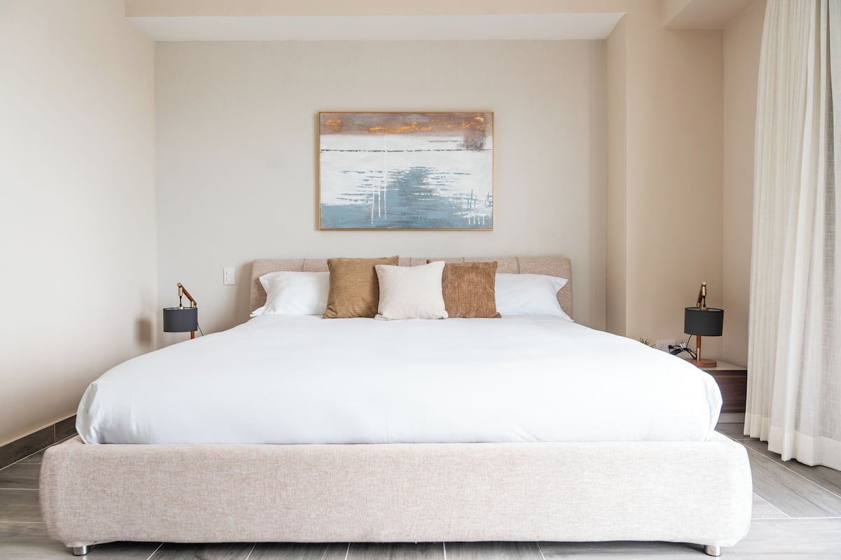 Una cama grande con ropa de cama blanca, un cabecero color beige y un cuadro en la pared. Hay dos mesitas de noche con lámparas.
