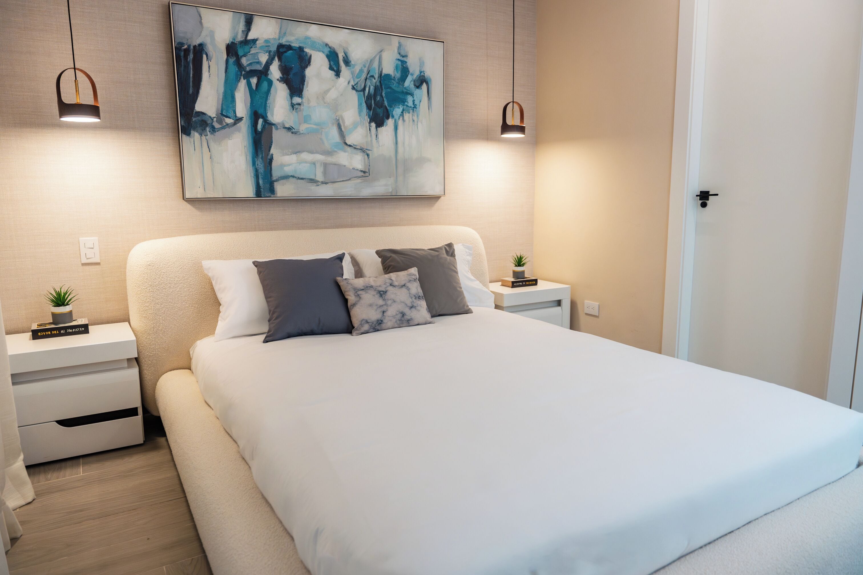 A modern bedroom with a large bed, two bedside tables with lamps, a wall-mounted abstract painting, and a door.