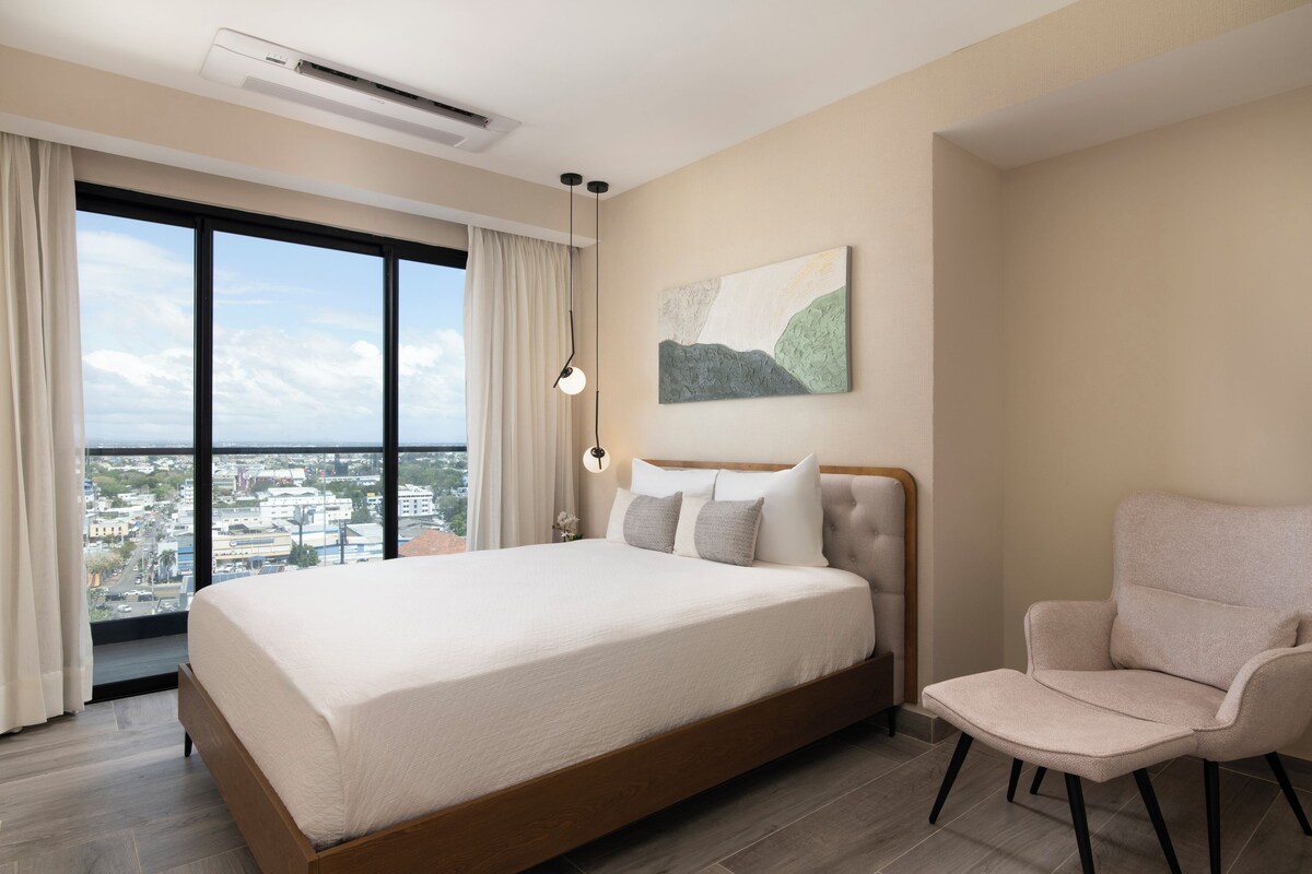 Ein modernes Hotelzimmer mit einem großen Bett, einem Sessel, Blick auf die Stadt und einem Bild an der Wand.