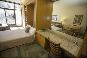 Ein kleines Hotelzimmer mit Bett, einem Schreibtisch, einem Stuhl, einem Esstisch und einem großen Fenster.