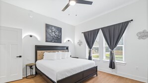A bedroom with a bed, a ceiling fan, a wall-mounted light, a window with curtains, and a small table. - Modern, Cozy and Spacious Home in Foley Alabama. (Foley)