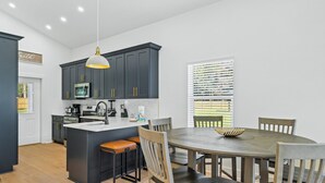 A modern kitchen with dark cabinetry, a central island, and a dining area with wooden chairs. - Modern, Cozy and Spacious Home in Foley Alabama. (Foley)