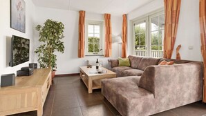 Ein Wohnzimmer mit einer Couch, einem Couchtisch und einer Pflanze. Es gibt Fenster mit Vorhängen, eine Lampe und Kunstwerke an der Wand.