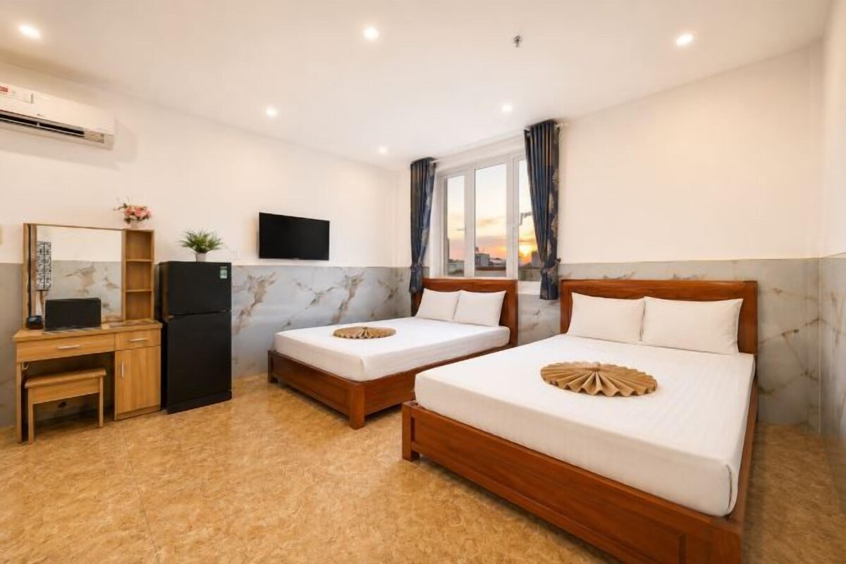 A hotel room with two beds, a desk, a refrigerator, a TV, and a window with curtains.