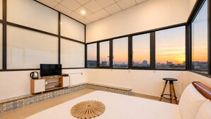 A bedroom with a bed, a TV, a small stool, and a view of the city at sunset.