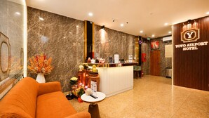 A hotel reception area with a marble wall, a reception desk, a wooden panel wall with a neon sign, and a plush orange sofa.