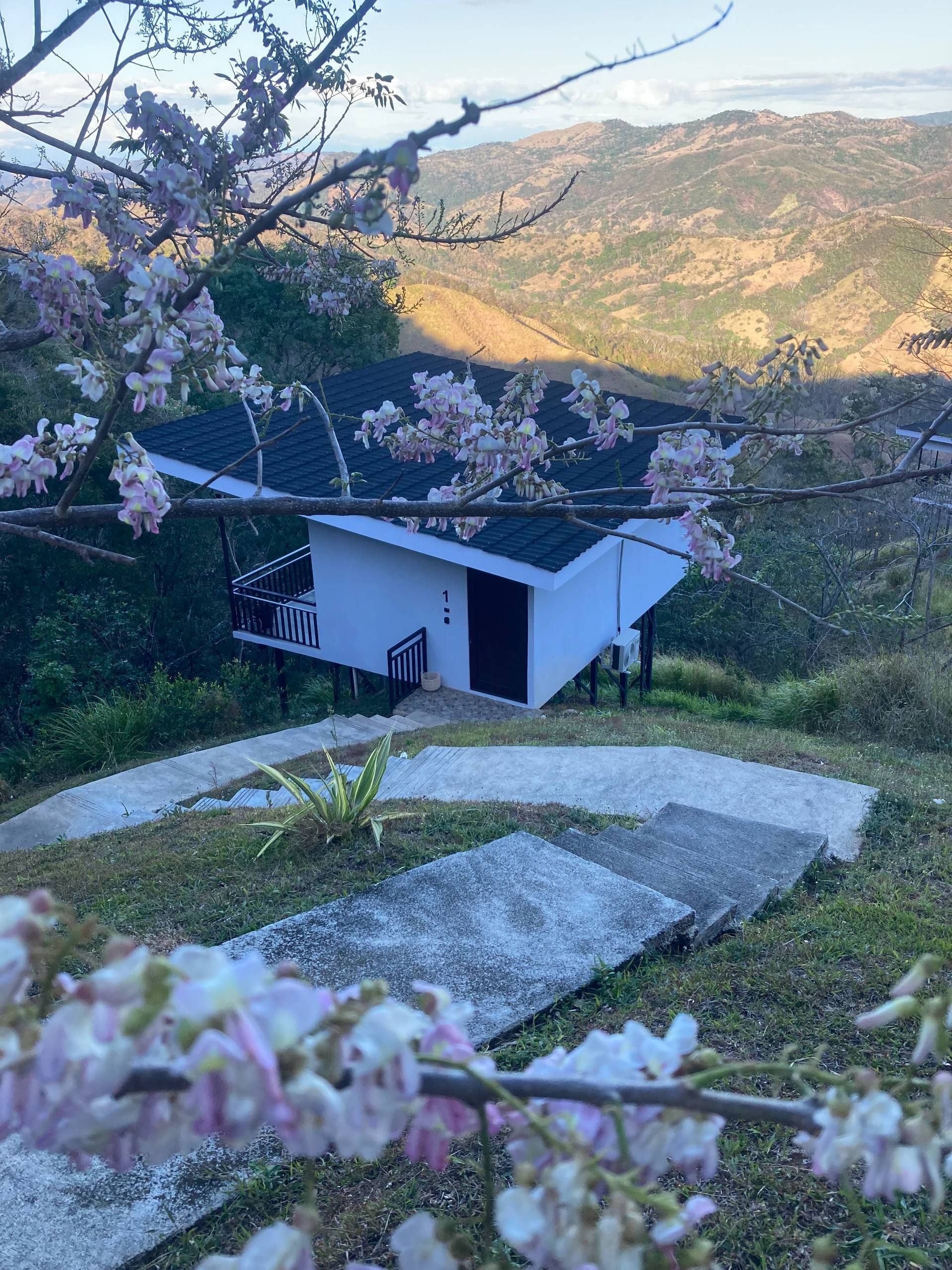A white building with a balcony, surrounded by a garden and a mountainous landscape.