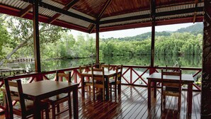 Un espacio al aire libre cubierto con mesas y sillas de madera, con vistas a un lago tranquilo y un bosque denso.