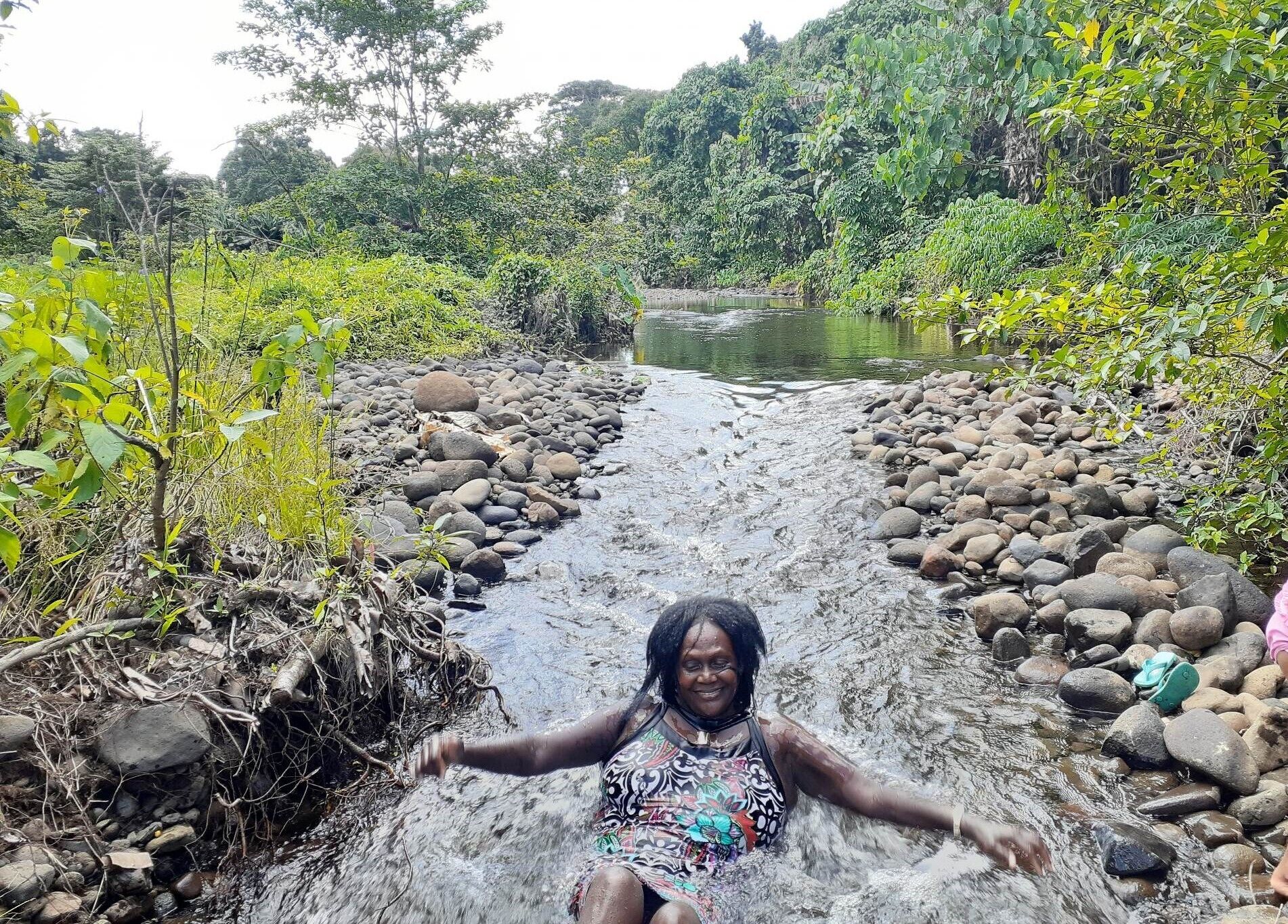 A woman sitting in a stream surrounded by rocks and greenery.