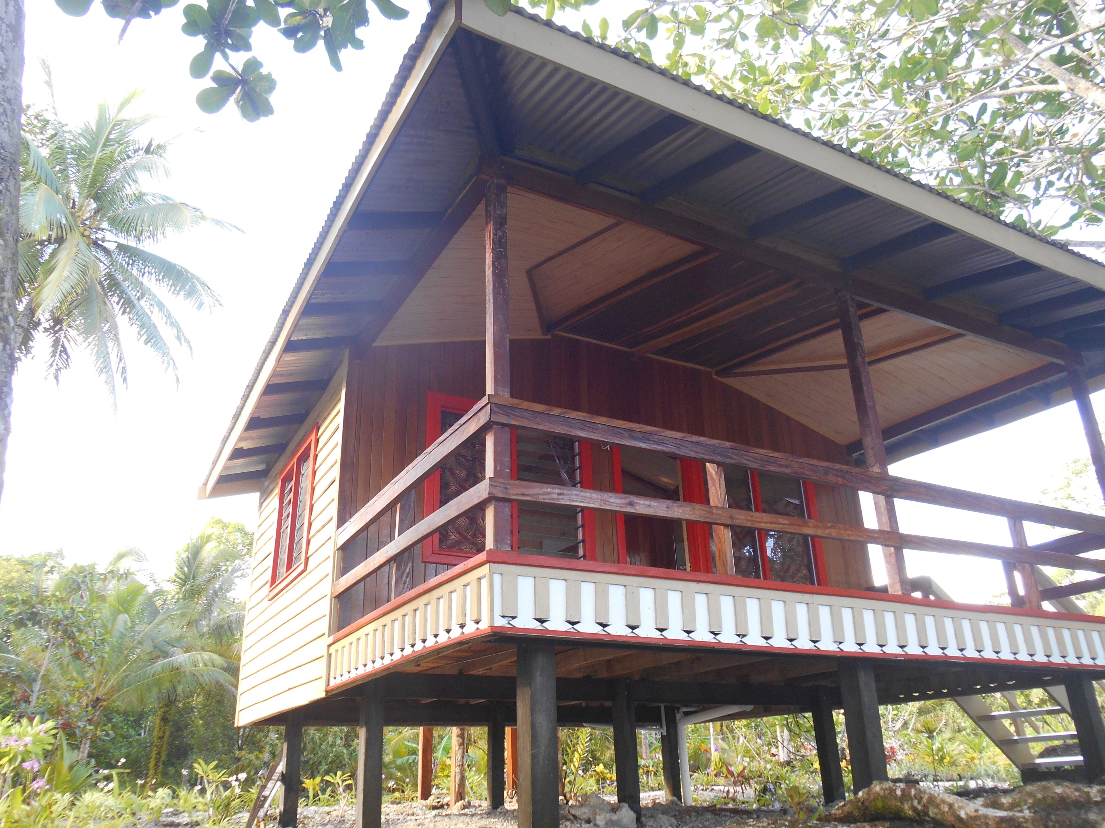 A wooden house with a balcony, surrounded by tropical vegetation.