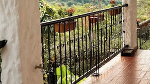 Un balcón techado con barandilla de metal, plantas en macetas y vistas a un paisaje verde exuberante.