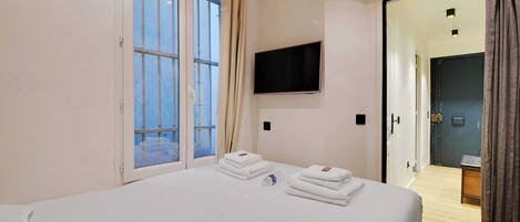 A hotel room with a bed, a TV mounted on the wall, a window with curtains, and a door leading to another room.