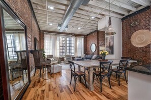 A spacious room with a wooden floor, a brick wall, a large window, and a dining area with chairs and a table. - Modern Stylish & Spacious Loft w/Indoor Parking (Birmingham)