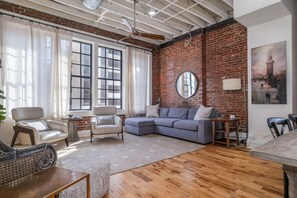 A living room with a brick wall, a large window, a sofa, two armchairs, a coffee table, and a painting on the wall. - Modern Stylish & Spacious Loft w/Indoor Parking (Birmingham)