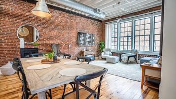 A spacious room with a brick wall, large windows, a wooden dining table, and comfortable seating.