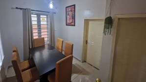 A dining room with a dark wooden table and six chairs, a window with blinds, a door with a green hanging ornament, and a painting on the wall. - Luxury Lagos House with 4 Bedrooms (Lagos)