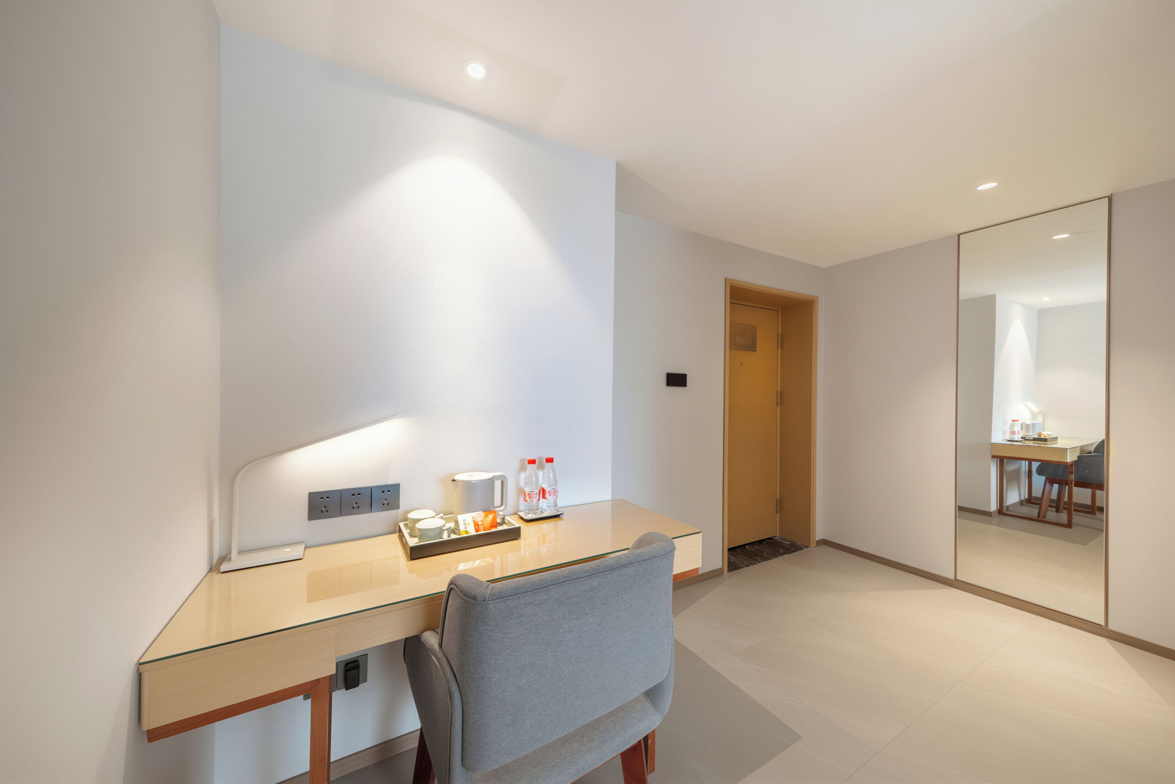 A modern hotel room with a glass desk, a chair, a small table with a tray of items, and a mirror on the wall.