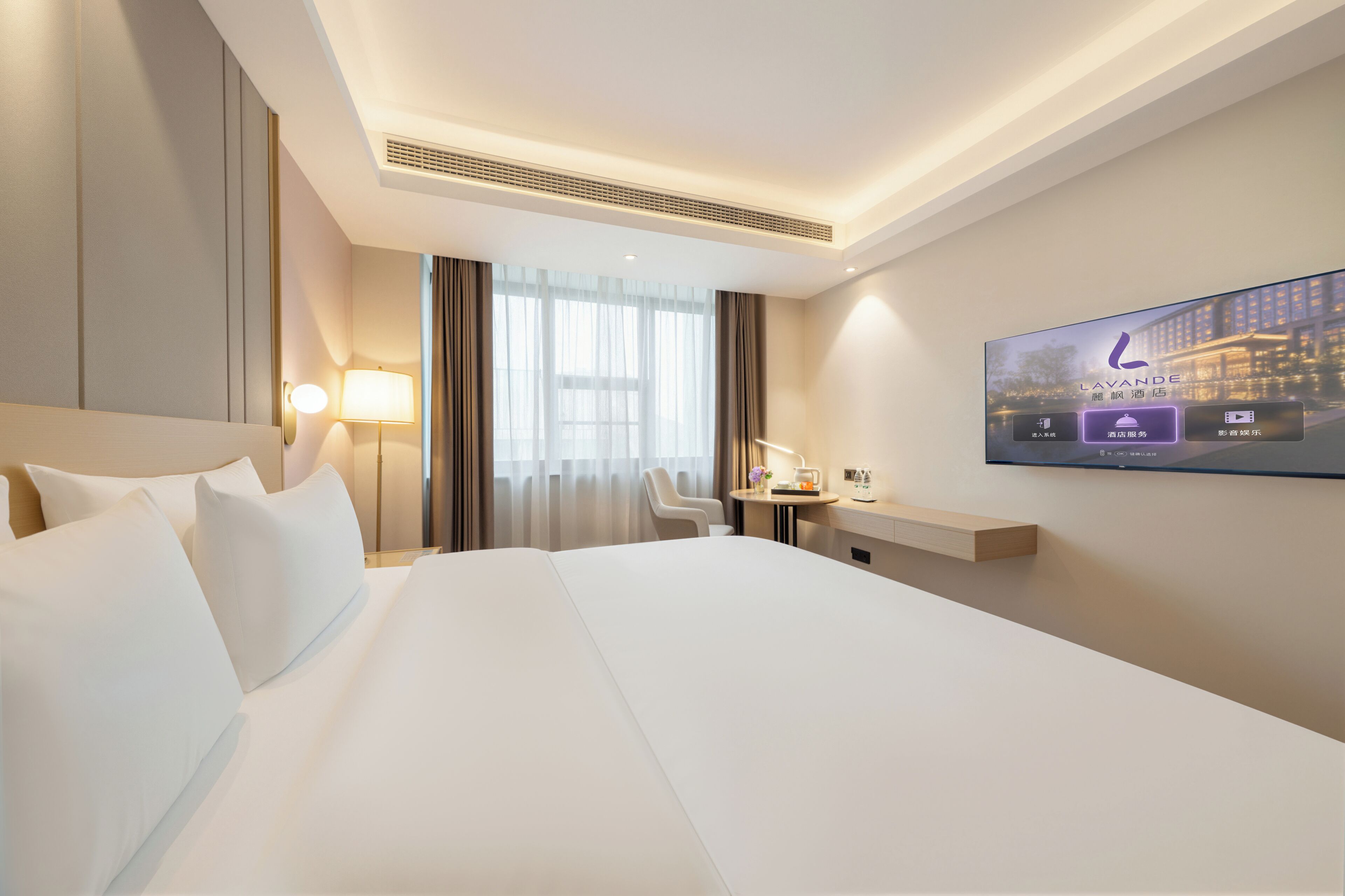 A hotel room with a large bed, a desk with a lamp, a TV on the wall, and a window with curtains.
