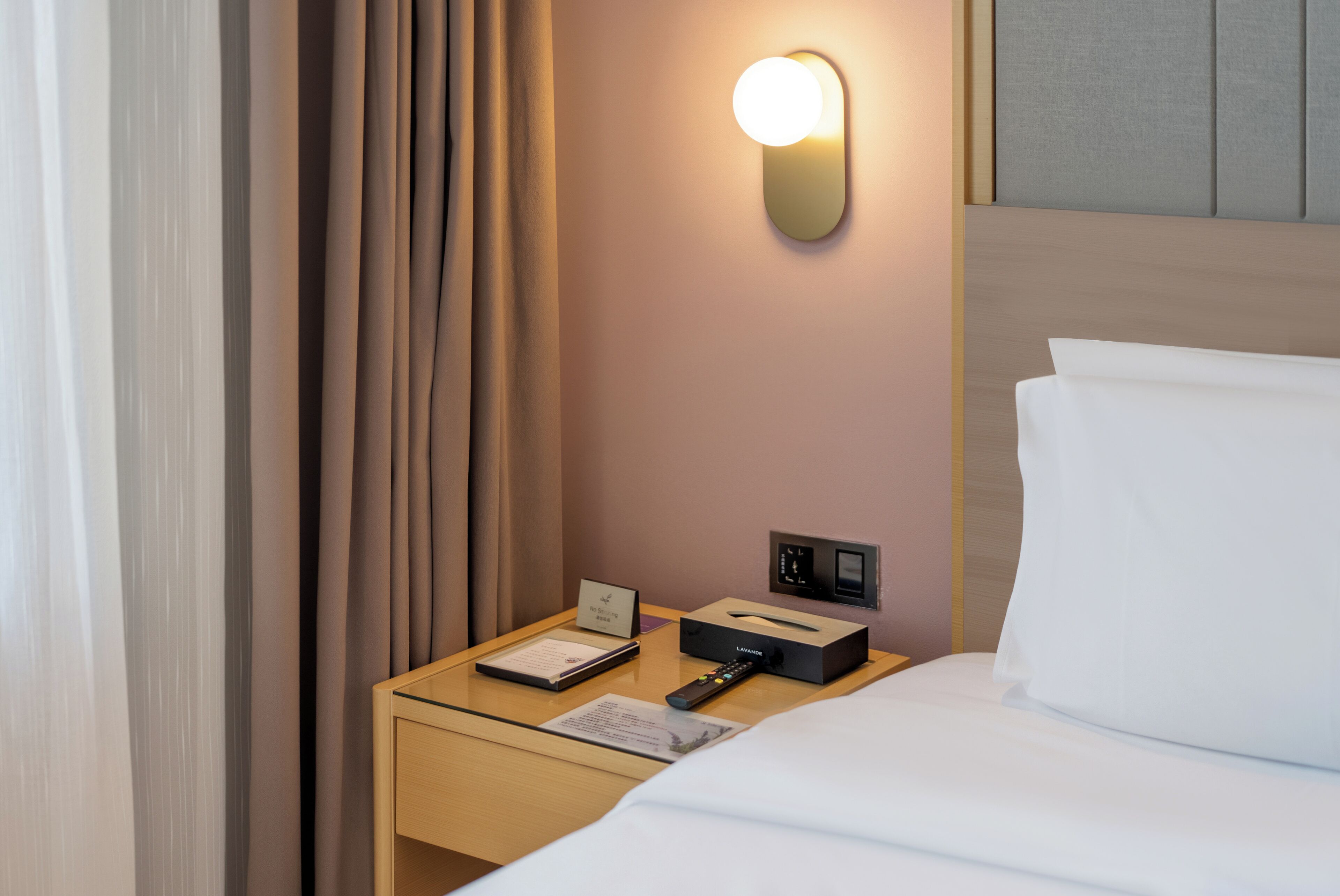 A hotel room with a neatly made bed, a bedside table with a remote control, a lamp, and a small card.