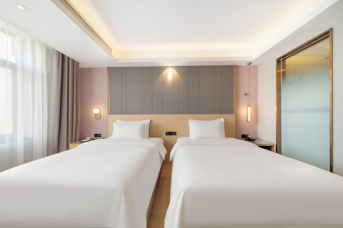 A hotel room with two beds, a nightstand with a phone, a wall-mounted light, and a large window with curtains.