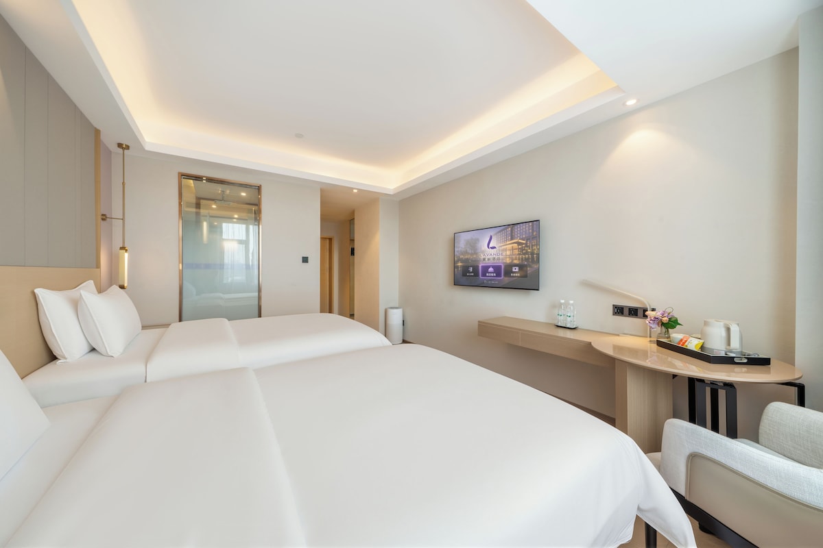 A hotel room with two beds, a TV, a desk with a lamp, and a small table with a flower vase.
