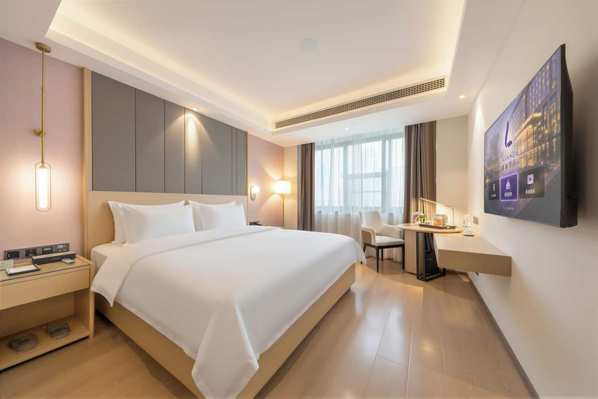 A modern hotel room with a large bed, bedside table, lamp, TV, and a desk with a chair.