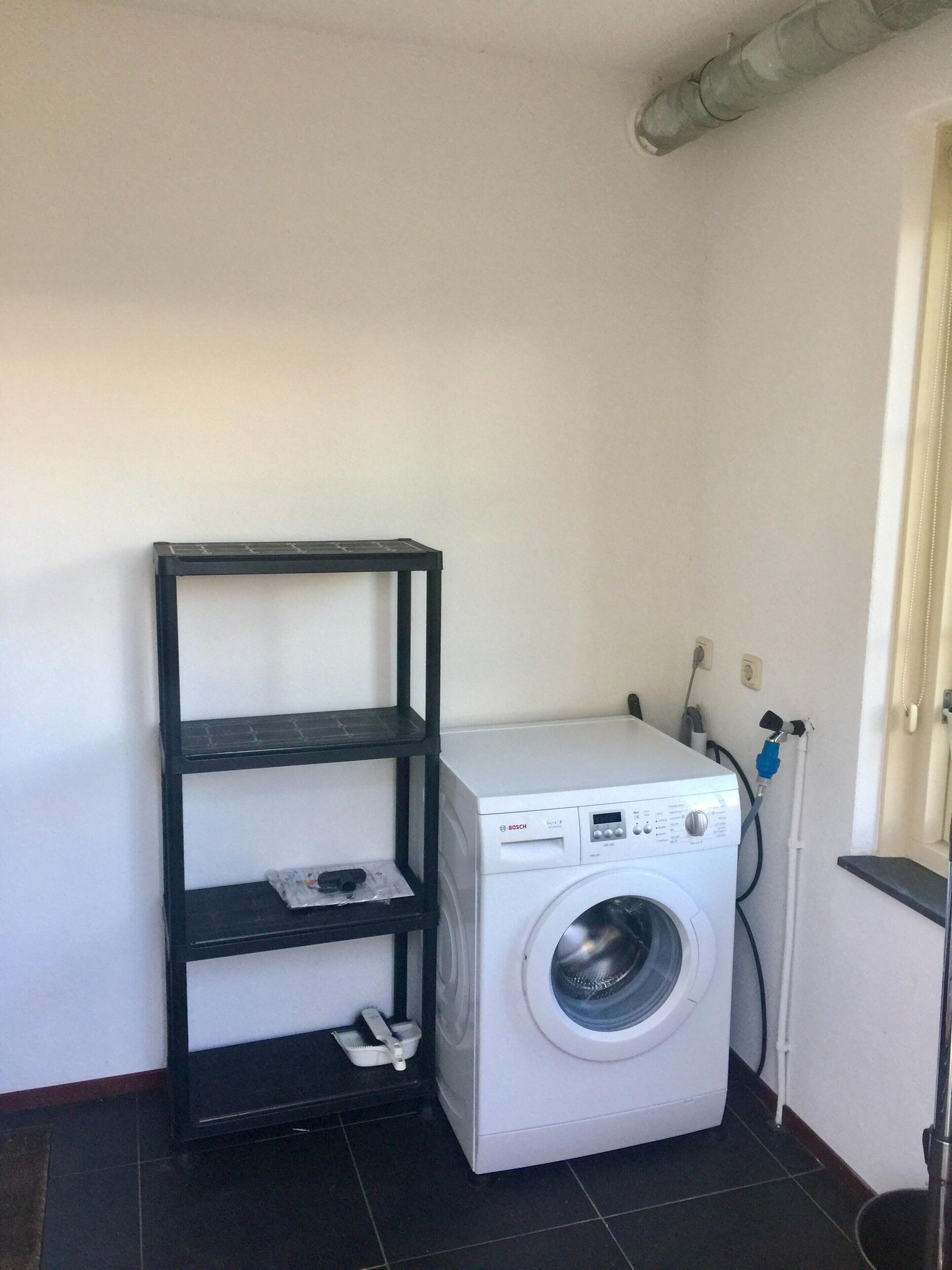 A compact laundry room with a Bosch washing machine, a black metal shelf, and a power outlet.